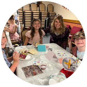 Two girls in dresses, an adult woman in a princess costume and a boy in a knight costume around a table with art supplies.