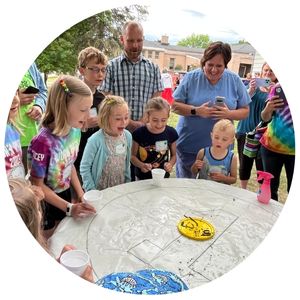 Kids cheering with adult behind them smiling around a table with a white tablecloth, yellow round lid with worms on it.