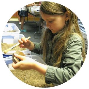 Picture of a girl with a brush in her hand as she brushes off sand from a relic out of the container with sand